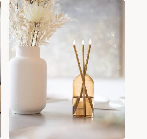 A Wylie Signature Set with gold candles in an amber glass vase on a table.